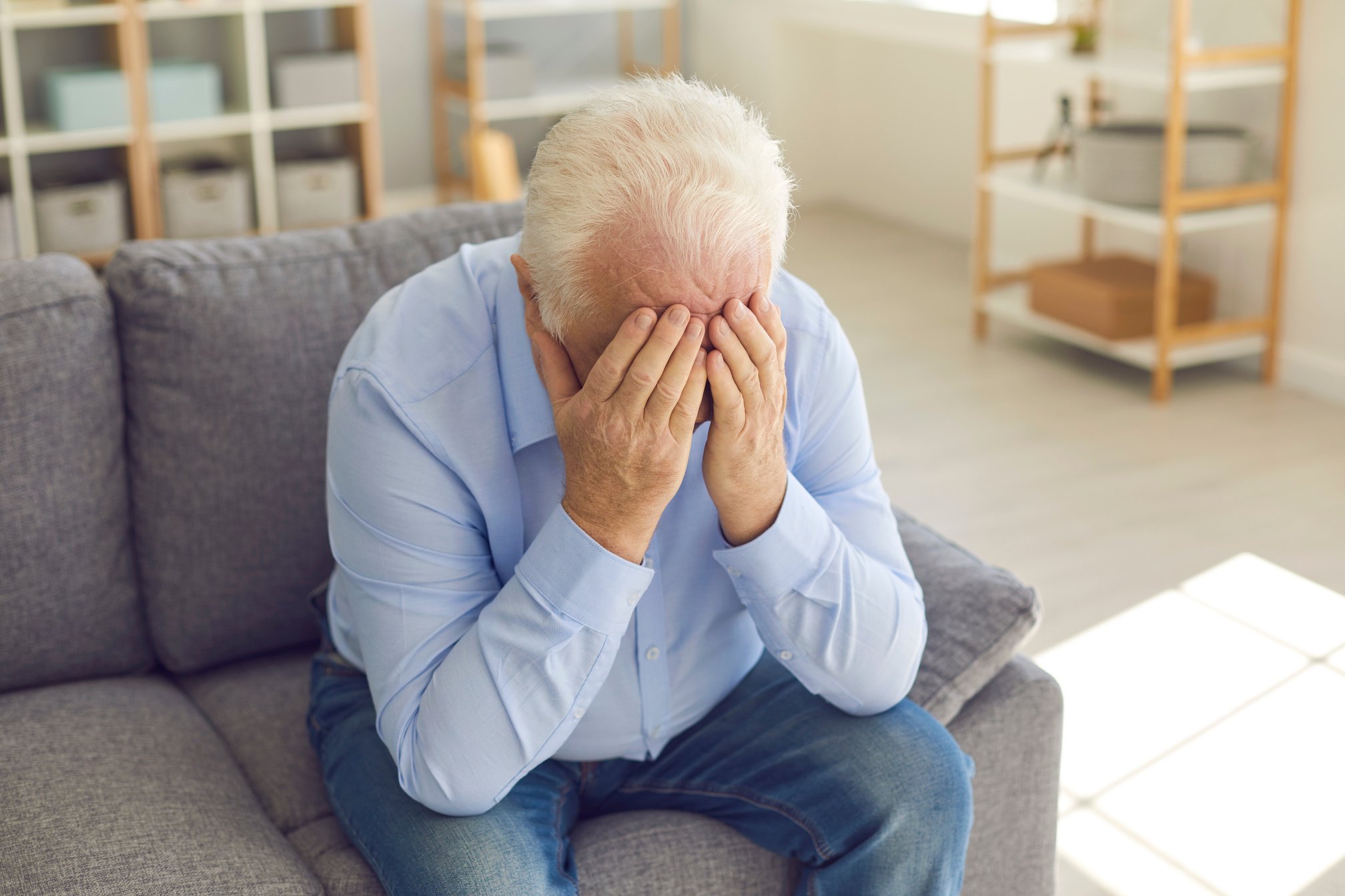 Shocked Senior Man Covers His Face with His Hands from Grief Sitting at Home on the Couch.